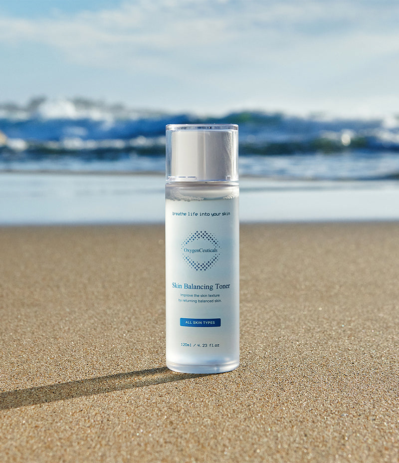 Clear toner-filled bottle of Skin Balancing Toner, for toning of skin, placed on the sandy shore.