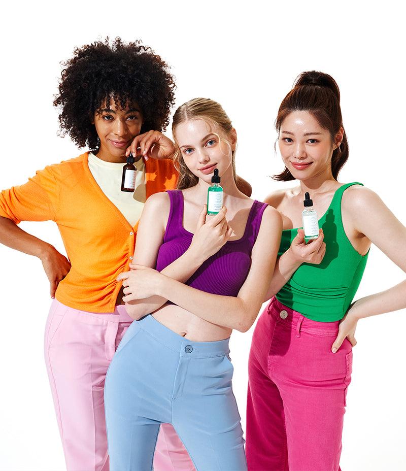 Three women stand smiling together with each holding a bottle of one of the Vitamin Serums.