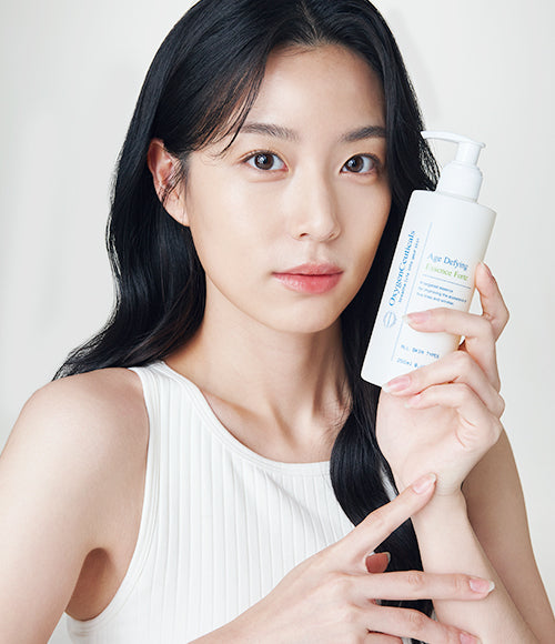 Woman holding up a bottle of Age Defying Essence Forte in front of a light gray backdrop.