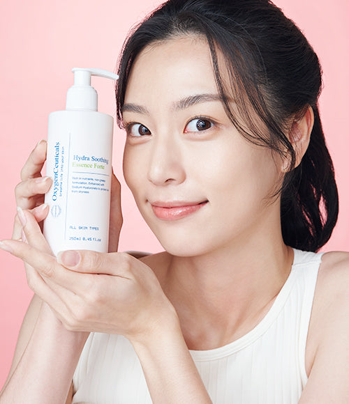 A happy woman holding a white bottle of Hydra Soothing Essence Forte in front of a pink background.