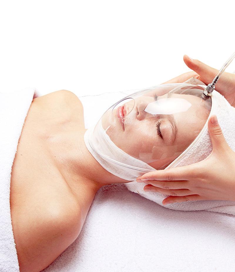 A woman undergoing a treatment, reclined on a table, with an oxygen mask from OxygenCeuticals OZ Portable.   Used for Oxygen facial. Can be used with masker oxygen, oxygen mask, oxygen serum, oxygen skincare, what is a misting device used for facial treatments, image oxygen facial.
