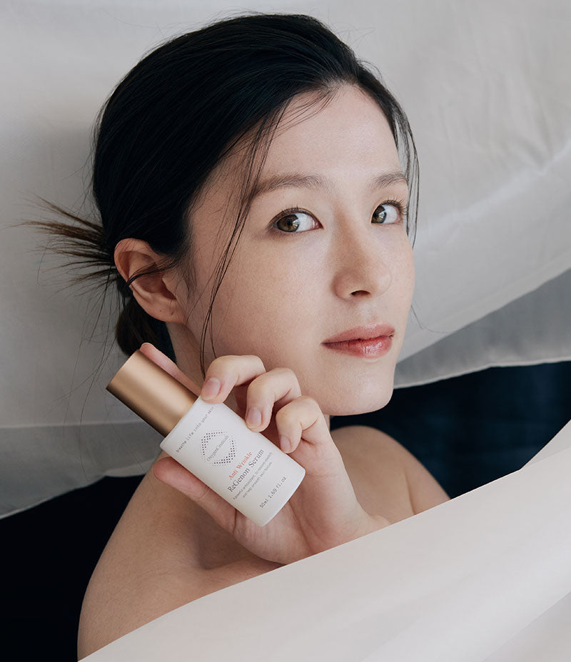  An elegant lady showcases a bottle of ReGenon Serum, a skin care product infused with Idebenone, known for its age-preventative properties.