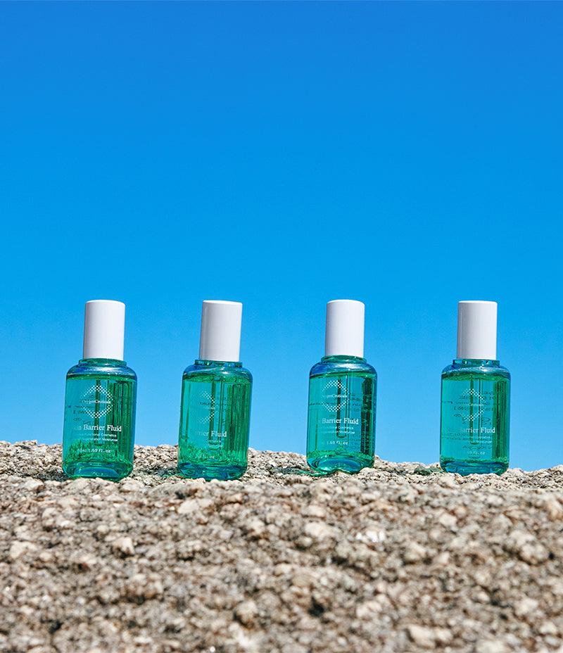 Collection of Skin Barrier Fluid bottles which improve skin elasticity, displayed on a rustic stone platform against the backdrop of a clear blue sky.