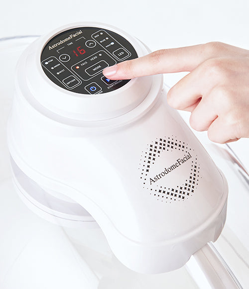 An individual using the control panel of a white machine, part of the OxygenCeuticals AstrodomeFacial LED skincare treatment device.